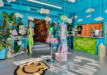 Colorful boutique interior with mannequins in vibrant outfits, hanging clouds, and a neon sign reading She Comes in Peace.