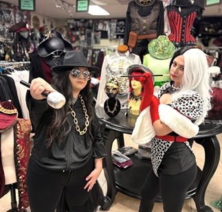 Two women in stylish black and white costumes pose confidently in a vibrant costume shop filled with hats and accessories.