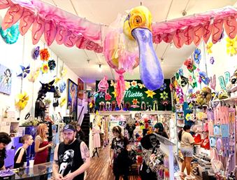 Colorful art shop interior with large pink bird sculpture, vibrant decorations, and shoppers browsing unique crafts.