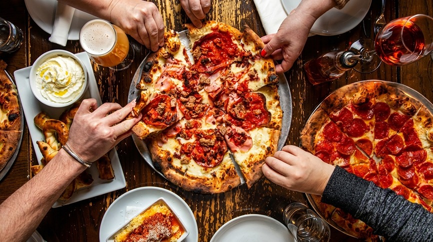 Hands reaching for slices of pepperoni and meat pizza on a rustic wooden table with drinks and sides