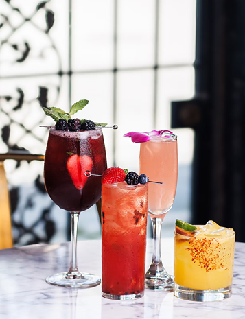 Four colorful cocktails garnished with berries, mint, and flowers on a marble table in a bright setting