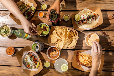 Overhead view of a wooden table with tacos, chips, dips, and drinks shared among people.