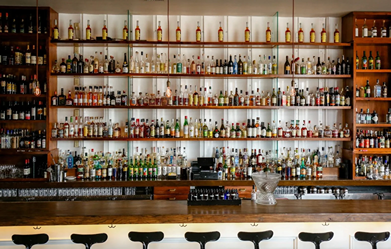 Well-stocked bar with various bottles of liquor on wooden shelves and black stools along the counter.