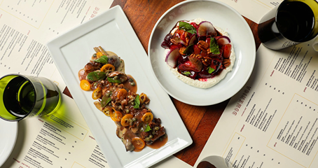 Gourmet dishes with sauces and fresh herbs served on white plates with menus and glasses on wooden table.