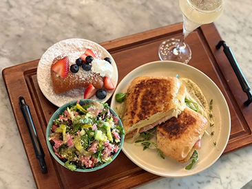 Grilled sandwich with salad, powdered sugar dessert with berries, and a glass of sparkling drink on a wooden tray.