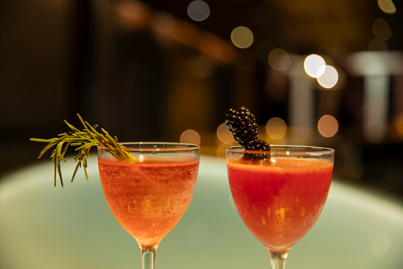 Two orange cocktails in glasses garnished with rosemary and blackberry, blurred warm bokeh background