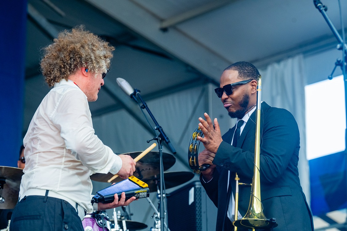 Musicians performing on stage with drums and trombone, one playing tambourine, under a tent roof.