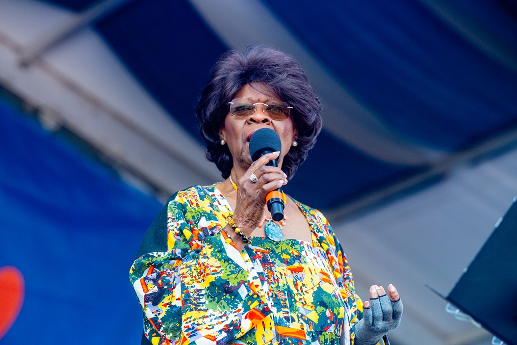Older woman with glasses speaking into a microphone on stage wearing a colorful floral blouse.