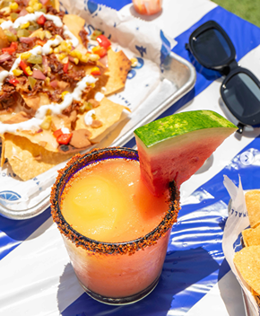 Frozen watermelon margarita with a chili rim and nachos topped with cheese and vegetables on a tray outdoors.