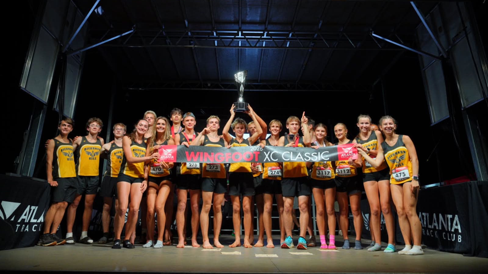 Youth cross country team in yellow and black uniforms holding a trophy and standing behind Wingfoot XC Classic banner
