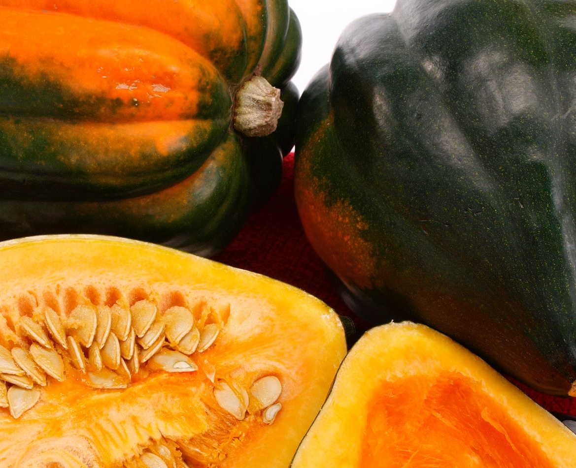 Acorn squash is place on a table. Two uncut acorn squashes set in the back (on top of the photo) and two halves of one acorn squash sit face up at the bottom of the photo.