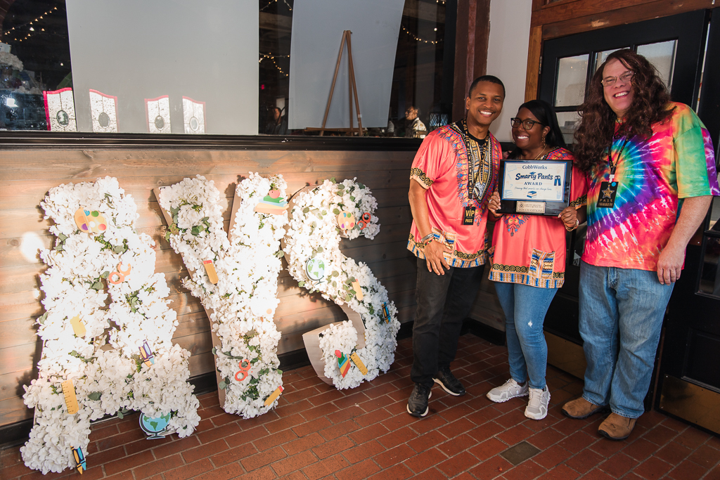 Three people smiling at an awards event with large floral letters AYS and a certificate on display.