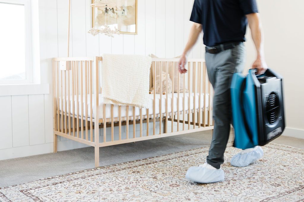 male Zerorez technician carrying a fan in a baby's room with a wooden crib and an oriental rug on a carpet 