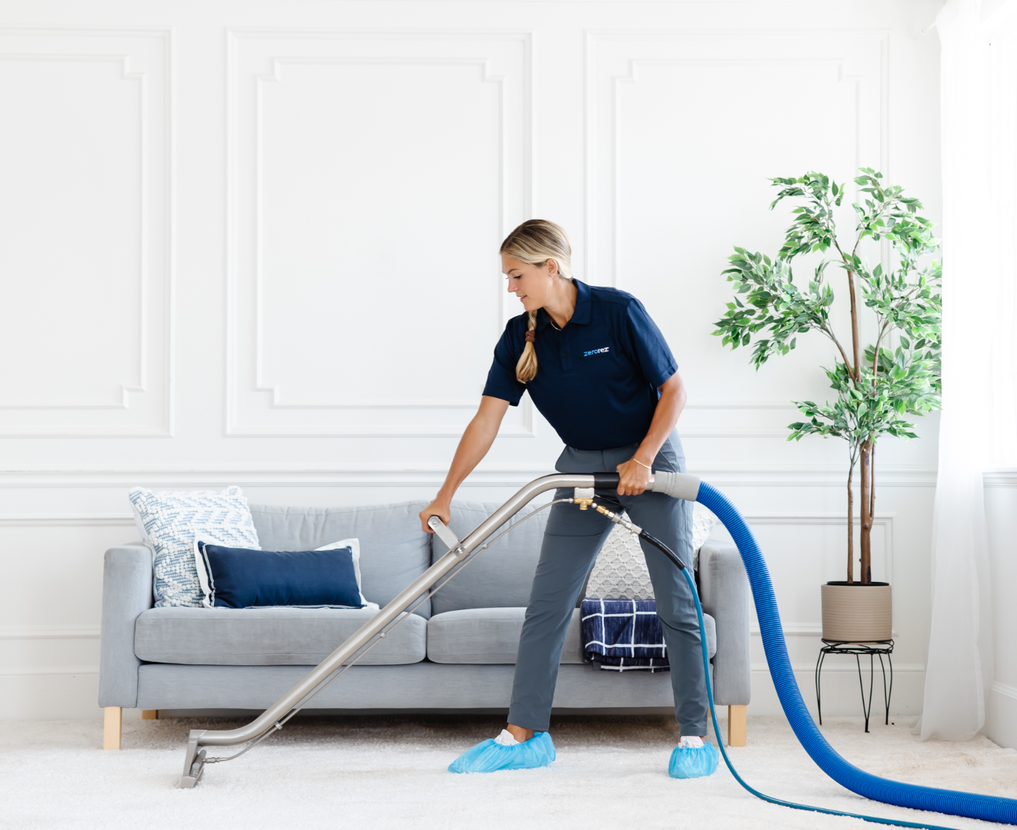 blond female Zerorez technician cleaning a white carpet in front of a gray couch in a living room before the house sells