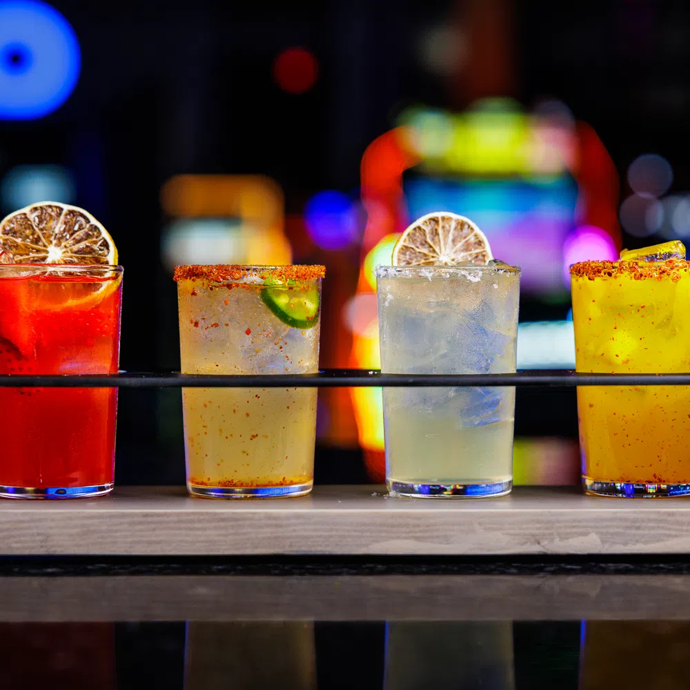 Four colorful cocktails with citrus garnishes served in a wooden flight holder on a reflective surface.