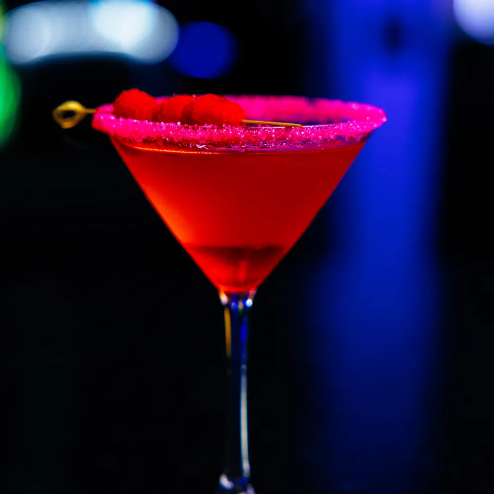 Red cocktail with sugared rim and raspberry garnish in a martini glass against a blurred neon background.