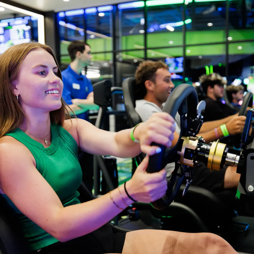 Young woman enjoys a racing simulator game with others in a vibrant, high-tech arcade setting.