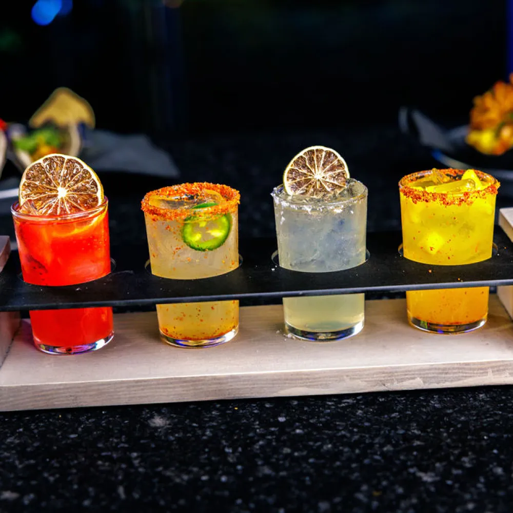 Four colorful cocktails garnished with dried lemon slices and spices on a wooden flight board.