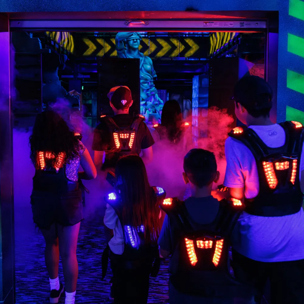 Group of people wearing glowing vests playing laser tag in a dark, foggy arena with neon lights.