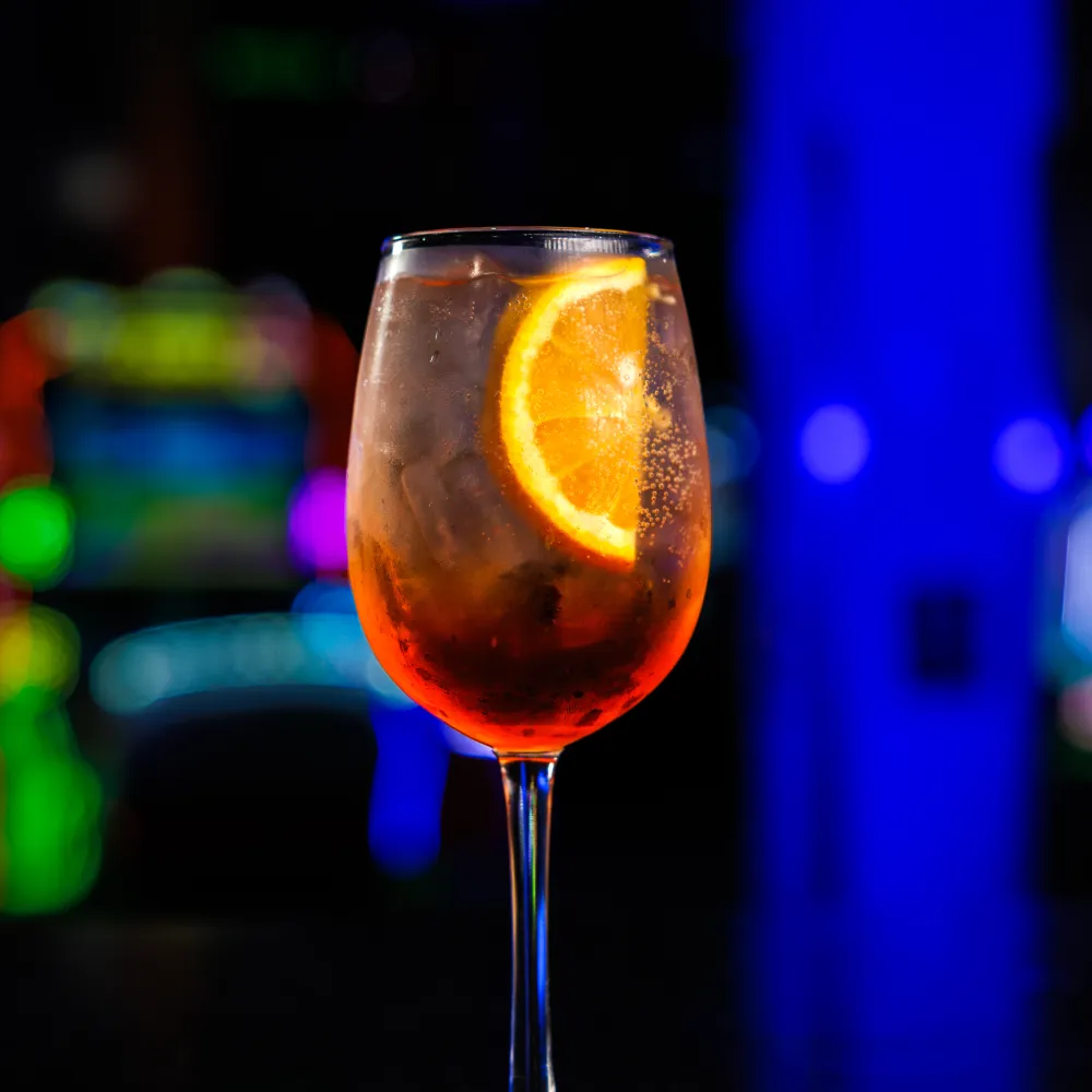 A chilled cocktail with an orange slice garnish in a wine glass against a colorful blurred nightclub background.