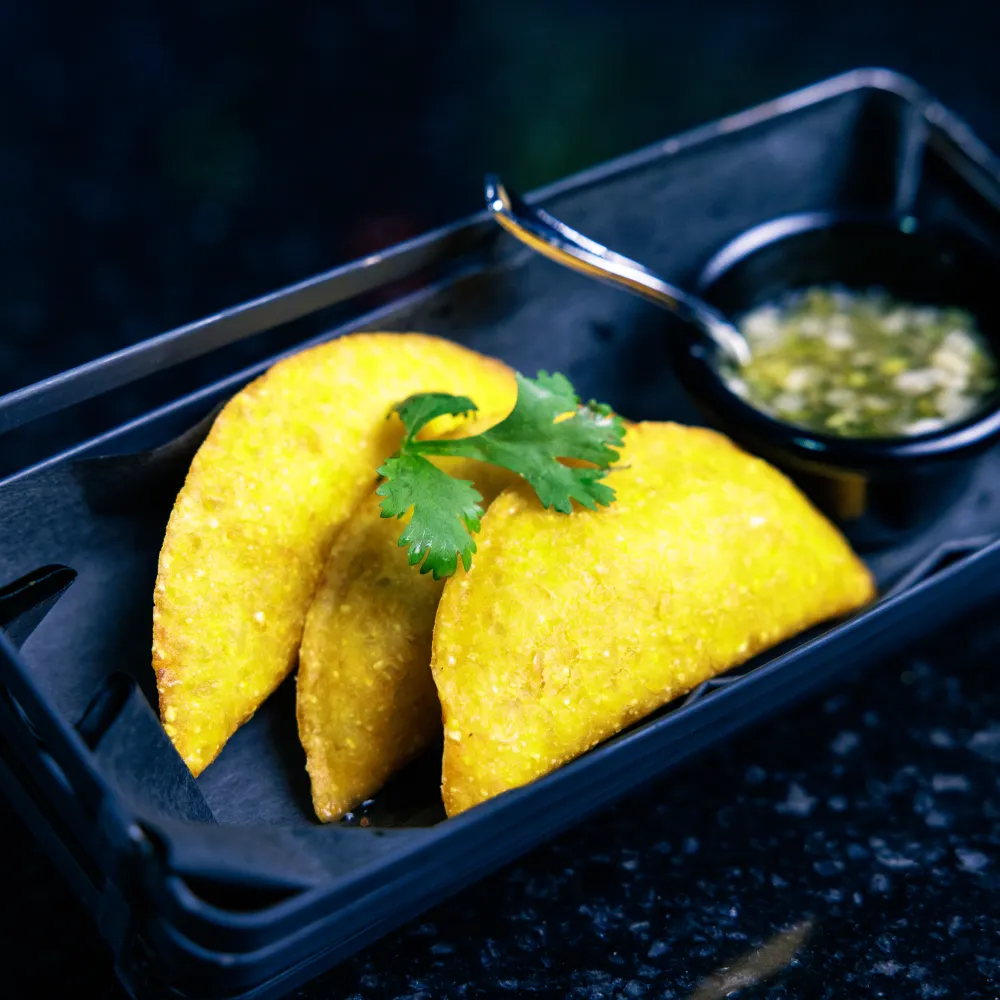 Golden fried empanadas garnished with cilantro served with green dipping sauce in a black tray.