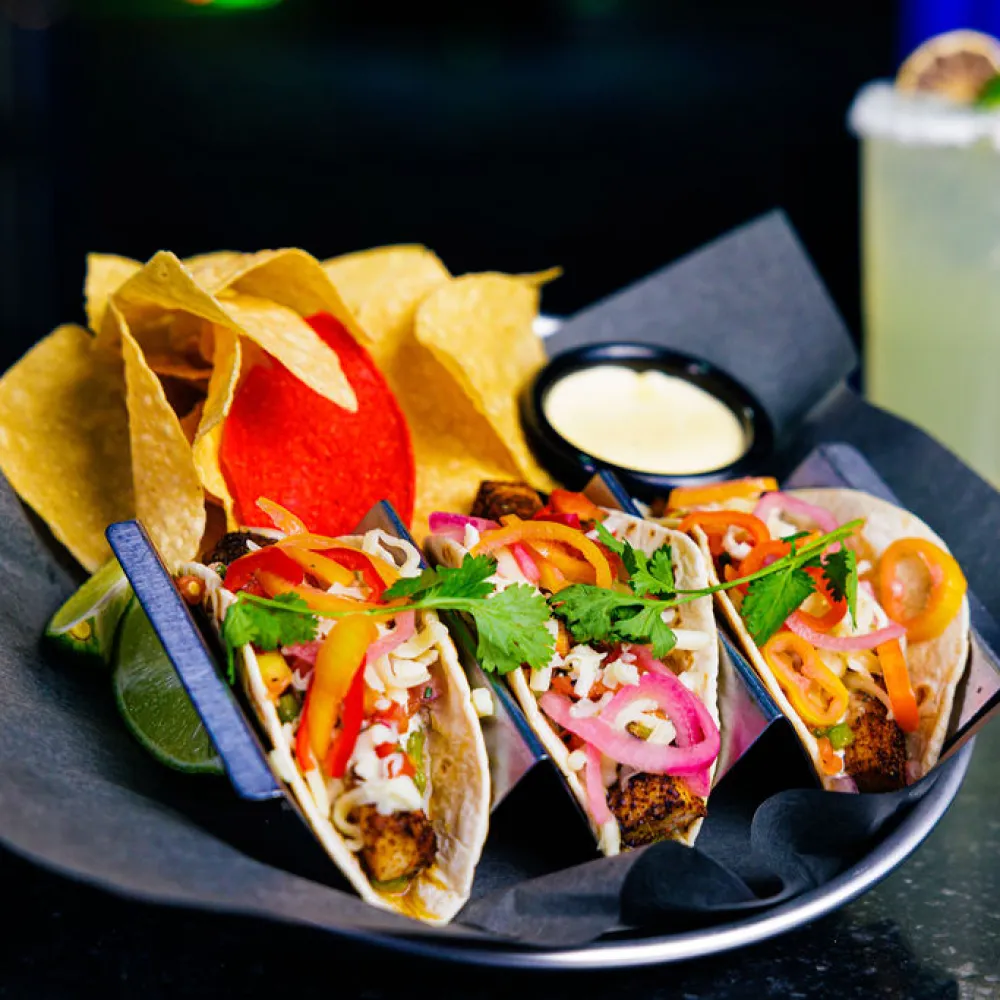 Plate of three tacos with grilled meat, pickled onions, peppers, cilantro, chips, queso dip, and a margarita glass.