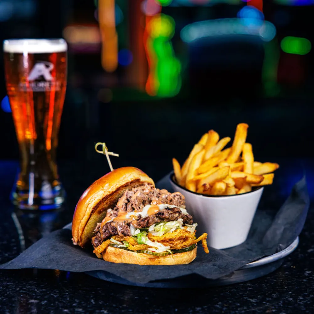 Double cheeseburger with fries and a glass of beer on a dark bar counter with colorful lights in the background.