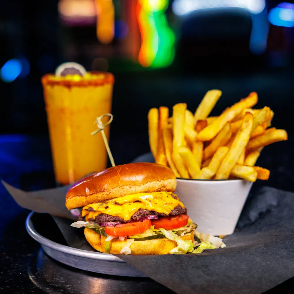 Cheeseburger with lettuce and tomato, cup of French fries, and a drink with colorful lights in background