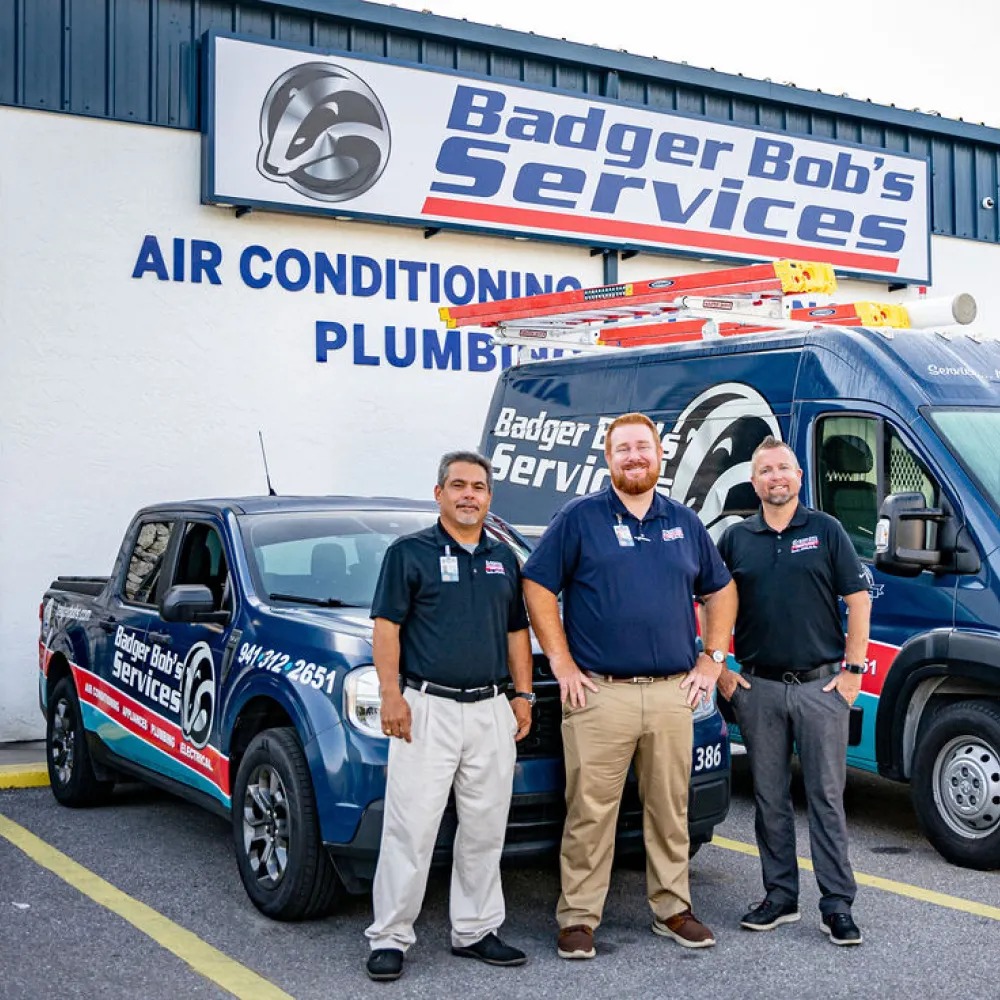 Three men stand in front of Badger Bob's Services trucks outside building advertising air conditioning and plumbing services.