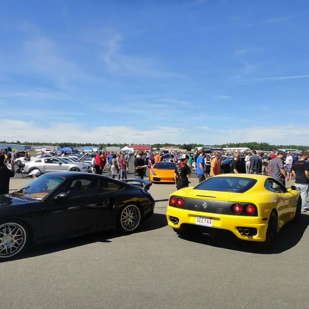 Car show with a black Porsche and yellow Ferrari surrounded by crowds under a clear blue sky on a sunny day