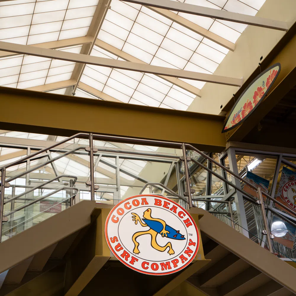 Cocoa Beach Surf Company logo sign hanging inside a modern building with glass railings and a skylight ceiling.
