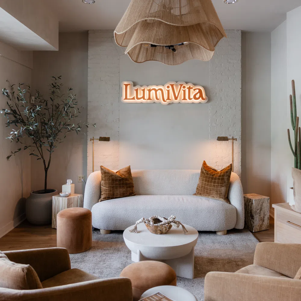 Cozy modern living room with white sofa, brown chairs, green plants, and LumiVita illuminated sign on wall.