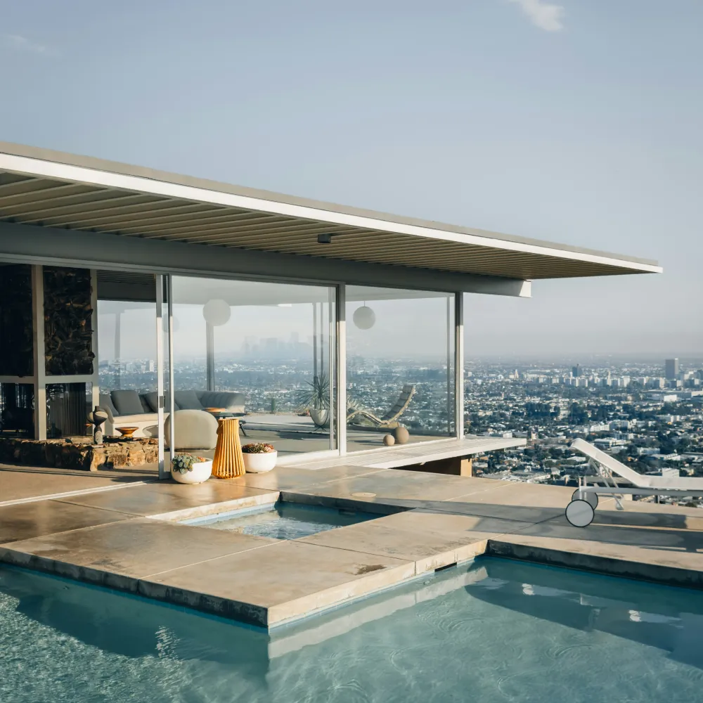 Modern house with glass walls overlooking a city skyline beside a clear blue swimming pool and hot tub.