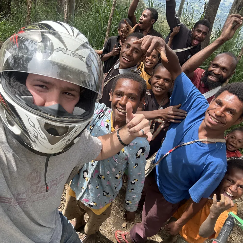 Group selfie of a person in a motorcycle helmet and local smiling people in a forested outdoor setting.
