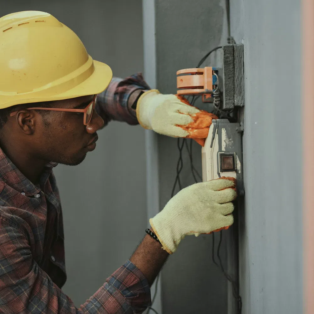 Electrician wearing a yellow helmet and gloves fixing an electrical panel on a grey wall indoors.