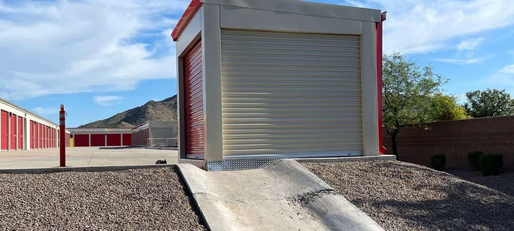Storage unit with closed shutter at the top of a concrete ramp surrounded by gravel under a blue sky.