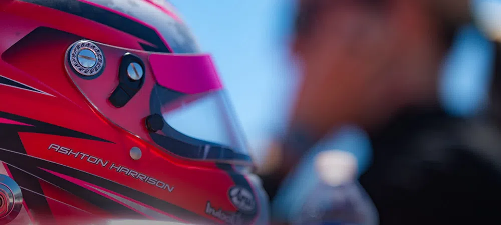 Close-up of a red racing helmet with Ashton Harrison name and blurred person in background on sunny day.