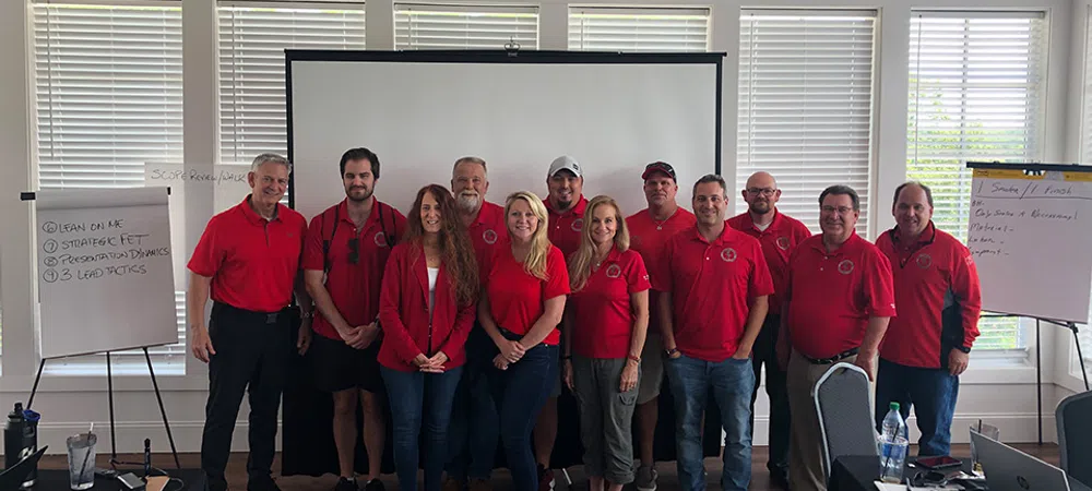 Group of professionals wearing matching red shirts posing in a conference room with presentation boards and projector screen.