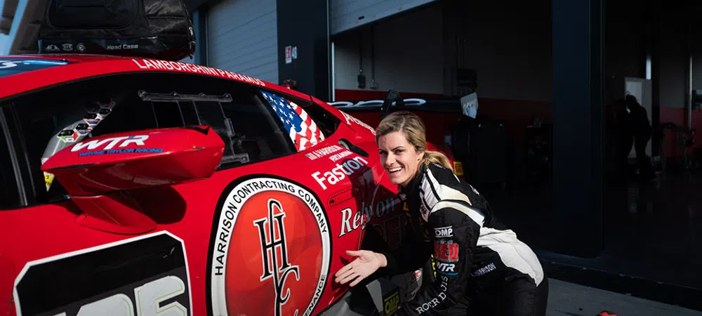 Female race car driver in black and white suit poses next to red number 125 Lamborghini race car in garage pit.
