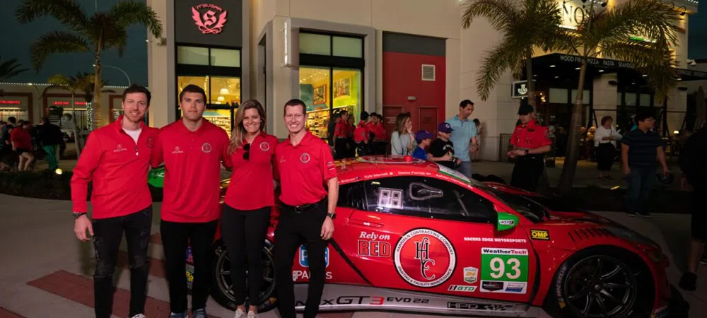 Four people in red shirts pose next to a red race car with sponsor logos outside a building at night.