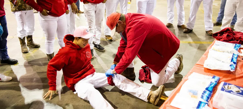 Man in red jacket and hat providing first aid to seated person in red hoodie and white pants during group event.