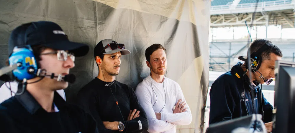 Four men in racing team gear monitoring screens and data in a motorsport pit area during daytime event.