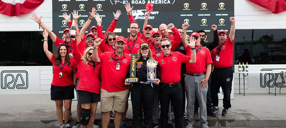 Racing team celebrating victory with trophy and champagne at Road America August 7-8, 2021 event podium.