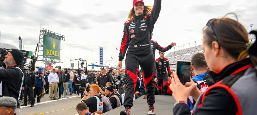Female race car driver in black and red suit waves to crowd at racetrack during event with spectators photographing her.
