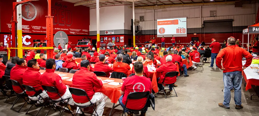 Large group of workers in red shirts attending a safety day meeting in a spacious industrial warehouse.
