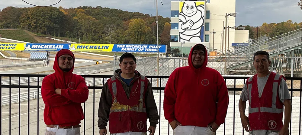 Four construction workers in red and white uniforms standing in front of a racetrack with Michelin ads and a mascot tower.