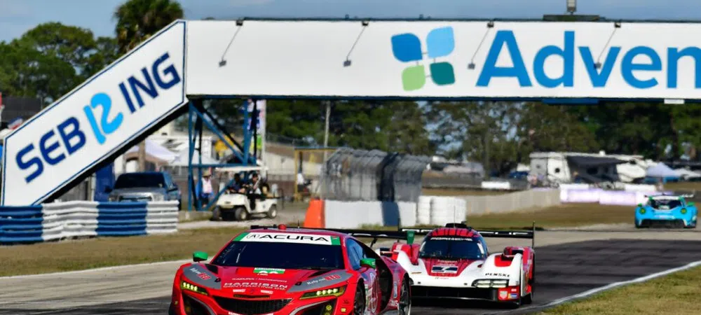 Red Acura race car leading a white and red prototype car on Sebring race track under blue sky.