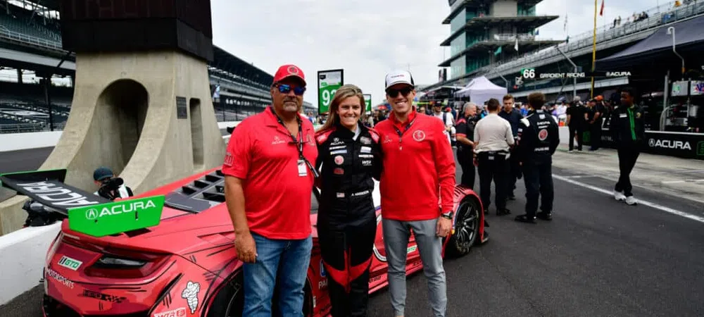 Three people in racing uniforms posing next to a red Acura race car at Indianapolis Motor Speedway racetrack.