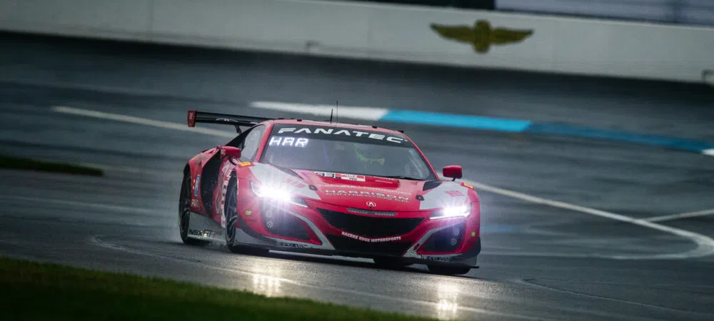 Red racing car with headlights on speeding on wet racetrack at night during motorsport event.