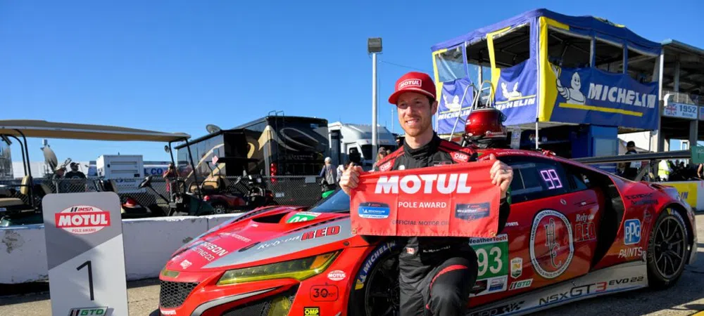 Race car driver kneels next to red NSX GT3 Evo holding Motul Pole Award banner at racetrack.